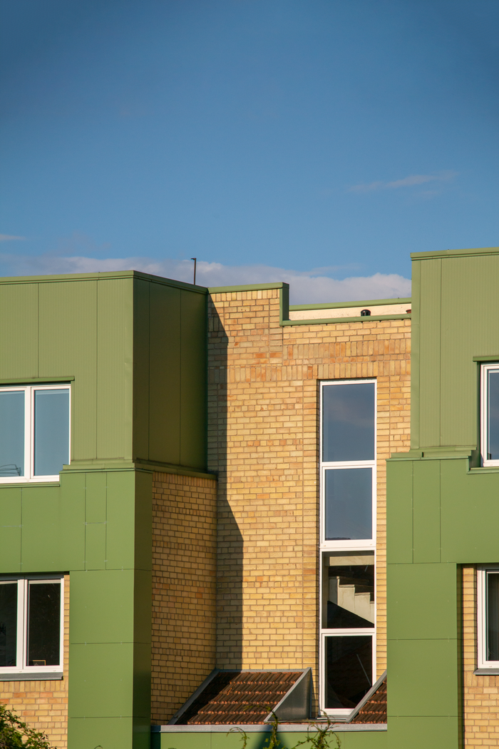 team-01 Sunlit modern building exterior with green and brick facade under clear blue sky.