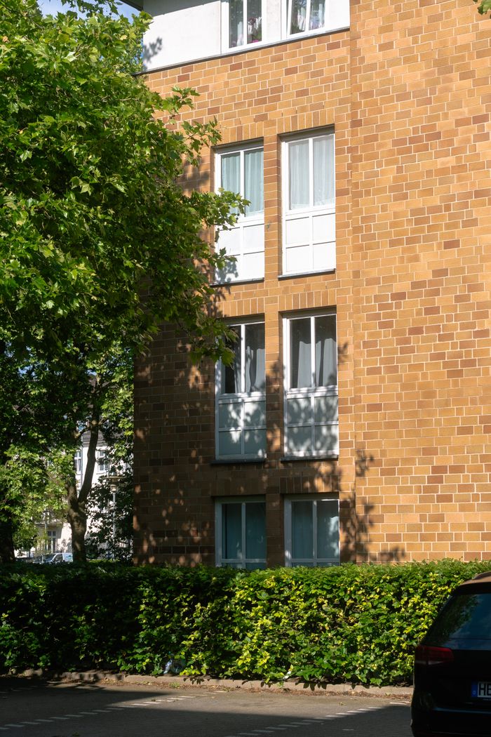 services-04 Sunlit brick apartment building facade with leaf shadows and greenery.